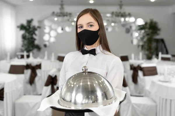 Female waiter in a protective medical mask holds a closed tray with a hot dish