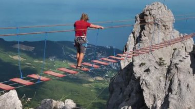 Genç bir adam dağın tepesine giden bir ip köprüden geçer.