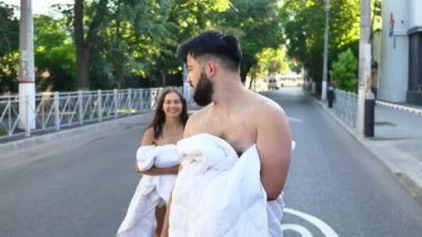 Beyaz battaniyeli bir adam ve bir kadın sabah güneşinde sokakta sarılıp öpüşürler.