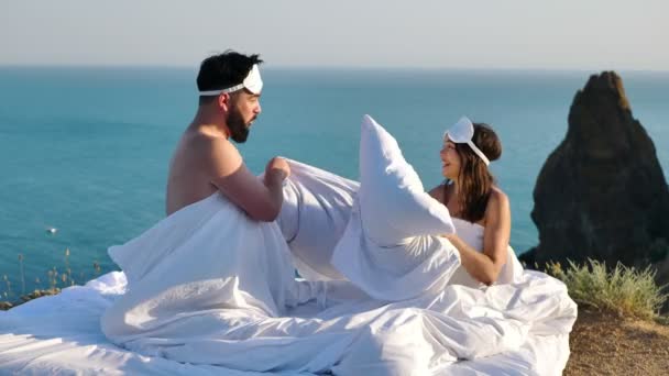 美丽的年轻夫妇在俯瞰大海的悬崖上 抱着枕头 躺在白色的床上打架 — 图库视频影像