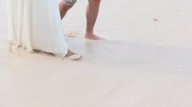 Yeni evlilerin balayı. Beyaz elbiseli çıplak ayaklı gelin ve romantik okyanus sahilinde yürüyen damat..
