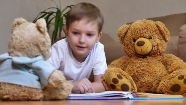 Щасливий Хлопчик Читає Книгу Пухнастих Іграшок Лежать Теплій Підлозі — стокове відео