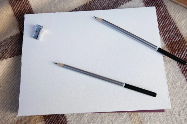 two gray graphite pencils and sharpener on a sheet of paper with a distinct texture. A sheet on a beige checkered blanket on a background of stack of books