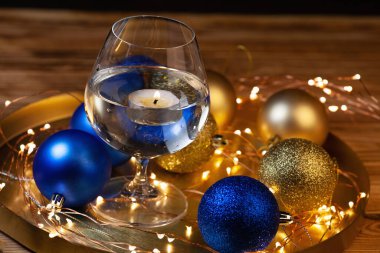 Parlak altın ve mavi topları olan Noel kartı, su bardağında mum ve Noel ışıkları