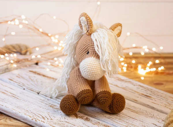 a wonderful crocheted soft toy. A figure of a brown horse with a lush white mane. On a wooden background behind bright led lights