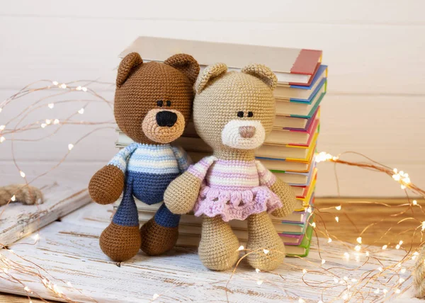 a pair of handmade knitted bears. A boy and a girl on a white wooden stand on the background of a stack of books. In the background are bright LED flashlights