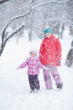 İki çocuk parkta kar altında dans ediyor ve gülüyor. Parlak kış kıyafetleri içinde küçük kız ve genç kız.
