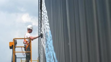 ALMATY, KAZAKHSTAN - 26 Ekim 2022: Miğferli genç adam endüstriyel beşik yan görünümlü püskürtücüyle depo cephesini boyuyor. 26 Ekim 'de Almaty' de dış duvara süs takmak