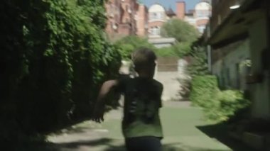 ALMATY, KAZAKHSTAN - AUGUST 22 2022: Blond boy runs along lane with wall covered with ivy on sunny day backside view. Little child plays on town street on August 22 in Almaty