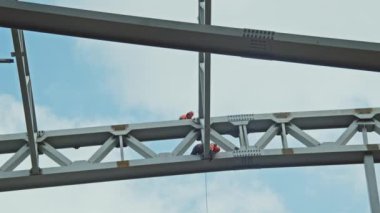 Builders work on metal carcass of sports stadium roof at construction site under cloudy sky. Dangerous operations