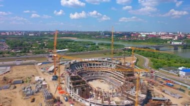 Sports center building carcass with tower cranes on riverbank against summer city bird eye view. Urban area development