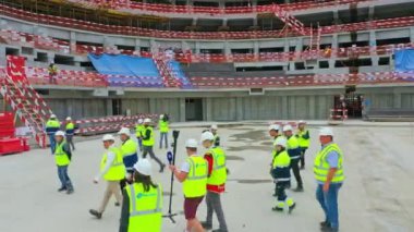Builders with shooting team in protective vests walk along construction site of stadium. Urban development presentation