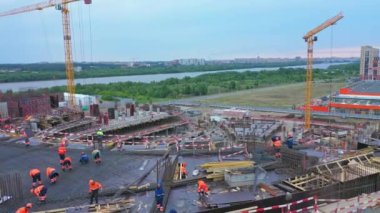 Builders in bright uniforms work on roof of future sports stadium at construction site bird eye view. Building industry