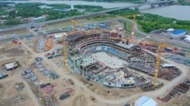 Sports stadium carcass with working cranes at construction site on riverbank bird eye view. Urban development technology