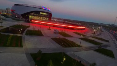 Young green park near contemporary sports arena building with bright neon illumination in evening town aerial view