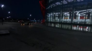 Truck cranes near glass facade of sports arena with large yard in night city first point view. Stadium building