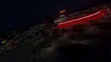 Athletic arena building with glowing neon lights and young park in night city first point view. Evening cityscape