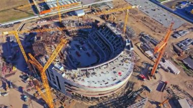 Atletizm sahasının şantiyesinde büyük vinçler ve ağır makineler çalışıyor. Tribünlerin hava manzaralı büyük beton stadyum kasesi.