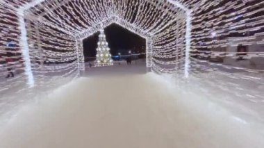 Decorations on snow in park with rink filled with people skating around Christmas tree. Festive spirit in park decorated with illumination