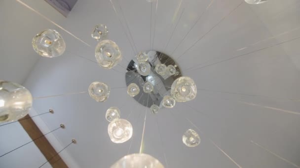 白い天井に掛けられた珍しい形のスタイリッシュなシャンデリアが家の前提を照らしています いくつかの電球は家の中のホールを照らす — ストック動画
