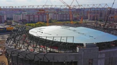 Sparkly roof of new constructed football arena against big city. Yellow building cranes constructing new stadium near town district at sunset