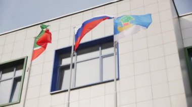 Okul binasının yakınındaki metal direklerde rüzgarın dalgalandırdığı bayraklar. Kentte açık gökyüzü altında ulusal sembollere sahip eğitim kurumları