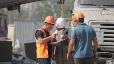 ALMATY, KAZAKHSTAN - 09 Mayıs 2023: Miğferli inşaatçılarla birlikte taskmaster kamyon yavaş çekimin yanında duran belgeleri kontrol ediyor. İşçi takımı inşaat alanında gazete okuyor.