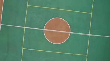 Spor salonunun yanındaki basketbol sahasında. Asfalt ve ağaçlarla çevrili beyaz işaretli boş yeşil spor sahası kameralar