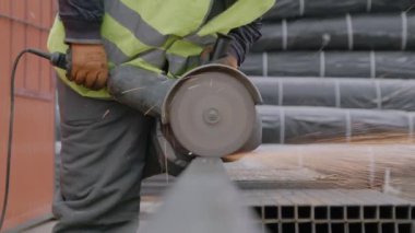 Güvenlik yeleği giyen işçiler inşaat alanında metal kalasları disk testeresiyle kesiyor. Çalışan dışarıda elektrikli aletle çelik boru kesiyor.