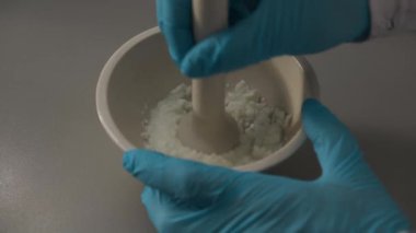 Lateks eldivenli laboratuvar asistanı, iş yerindeki havanda beyaz taneli malzeme eziyor. Uzman testler için madde örneği hazırlıyor.