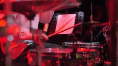Müzisyen, festival konserinin yakın çekimlerinde bateri setinde rock müzik çalar. Yetenekli davulcu kırmızı ışıklarda sahnede ahşap sopalarla hızlı davul çalar.