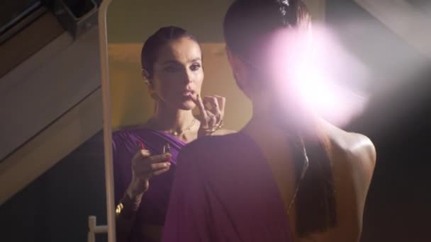 アルマニー カザフスタン 2024 夜のドレスの魅力的な女性は 鏡の前に指で口紅を適用します 若い女性はドレッシングルームでのパフォーマンスのために準備します — ストック動画