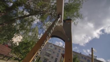 Demir salıncak zinciri yaz günü güneş ışığında parlar. Rüzgarda yavaşça sallanırken arka planda çok katlı bir apartman var.