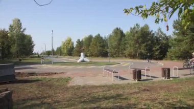 Park alanında banklar ve ortasında beton döşeme var. Tatilciler bisiklet sürerler ve park alanında dolaşırlar.