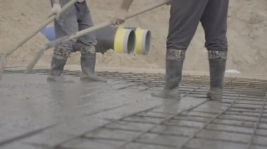 Erkek işçiler inşaat alanında havan topu seviyesinde çalışıyor. Kürekli adamlar ağır çekimde beton harç ağlarına döküyorlar.