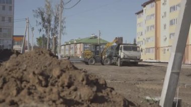 Kazıcı, çöp kamyonunun arkasına boru hattı hendeği kazdıktan sonra kalan toprağı boşaltıyor. İşçilerin rahatlığı için inşaat alanının boşaltılması