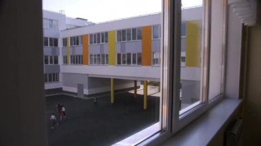 Sınıfın geniş penceresi okul bahçesini büyüleyici bir şekilde gösteriyor. Üniformalı öğrenciler okula yürür ve gün için planları tartışırlar.