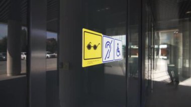 Engelliler için girişe uyarı levhası asılmış. Etiketler koyu cam kapıyla keskin bir tezat oluşturuyor. Modern binanın şık kapısı.