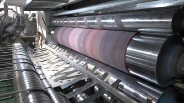Düz kağıt parçaları makinede silindirlerin altında hareket ediyor. Mekanizma hammaddeyi işlemek ve karton oluşturmak için atölyeye taşıyor