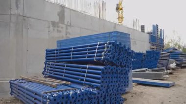 Mavi metal destekler kabloyla bağlanmış ve istiflenmiş. Temelinde takviye olan uzun beton duvar ve vinç yükseliyor.