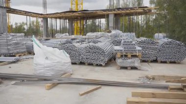 Metal borular ve inşaat malzemeleri yerde yatıyor. Beton sütunlu kısmen inşa edilmiş yapı, temelde vinç altında duruyor