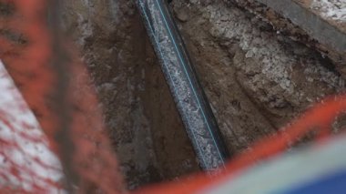 İnşaat için siper içine boru döşenmiş. Siyah, mavi şeritli boru. Sert toprak duvarlarla çevrili. Yeraltı tesisatını işaret ediyor.