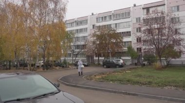 Park etmiş arabalarla apartman bloğu ve yolda yürüyen yaya bir kadın. Sonbahar ağaçları ve çimlerin olduğu bir alan sakin bir yerleşim yeri hissi veriyor.