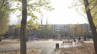 Sonbahar günü ağaçlar ve banklarla çevrili boş bir oyun alanı ve yere saçılmış yapraklar. Güneşli havada konut binaları ve arabalar