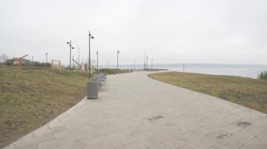 Bulutlu gökyüzünün altındaki su kütlesine giden yol. Alanda ahşap kule ve kamu parkında kaldırım boyunca sıralı metal banklar yer alıyor.