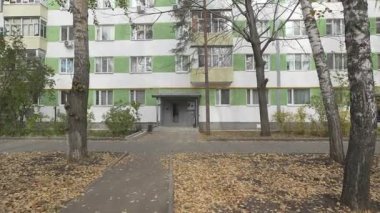 Sonbaharda ağaçlar ve düşen yapraklarla çevrili bir binanın girişinde. Tekrarlanan pencereler ve doğa elementleri sakin bir atmosfer yaratır