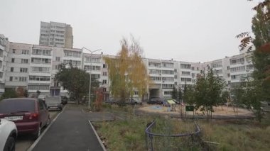 Ortasında küçük bir oyun alanı olan bir apartman dairesi. Sonbahar yapraklı ağaçlar ve park etmiş arabalar sessiz bir mahalle ortamı yaratıyor.