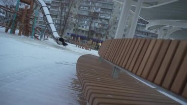 Pavilion bankında karla kaplı oyun parkı huzurlu bir görünüm sunuyor. Yapı, çocuklar için tasarlanmış açık oyun alanını çerçevelerken barınak sağlıyor