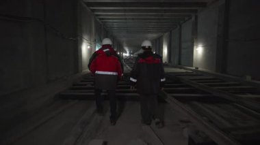 İşçiler inşaat halindeki loş metro tünelinde yürürler. Güvenlik teçhizatı karanlık çevreyle çelişiyor. Tünel çiğ ve endüstriyel
