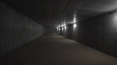 Loş ışıklandırma yayaları dar alt geçitten geçirir. Geçit metro platformlarına açılıyor. Yerin derinliklerinde bastırılmış bir ton yaratıyor.